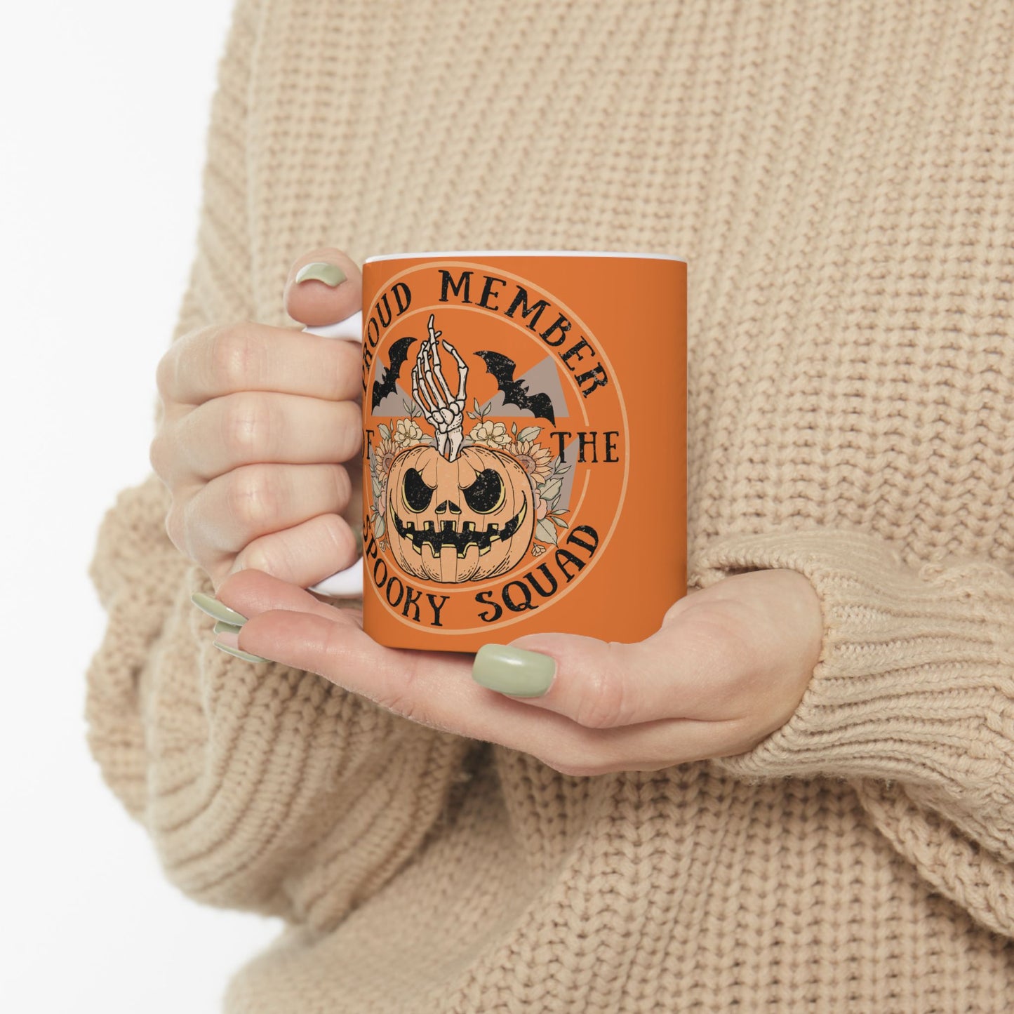 Halloween ceramic mug with a skeleton hand coming out of a jack o lantern with flowers behind it, two bats above it, surrounded by the words "proud member of the spooky squad" in a circle, orange background.