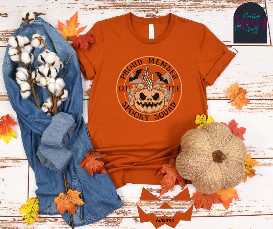 Pumpkin Halloween shirt with jack o' lantern in front of sunflowers, a skeleton hand popping out the top, two bats above, and the words "proud member of the spooky squad" making a circle around it all, faded vintage look, in autumn.