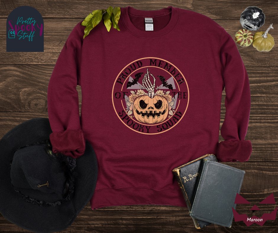 Halloween sweatshirt with a skeleton hand coming out of a jack o lantern with flowers behind it, two bats above it, surrounded by the words "proud member of the spooky squad" in a circle, in maroon.