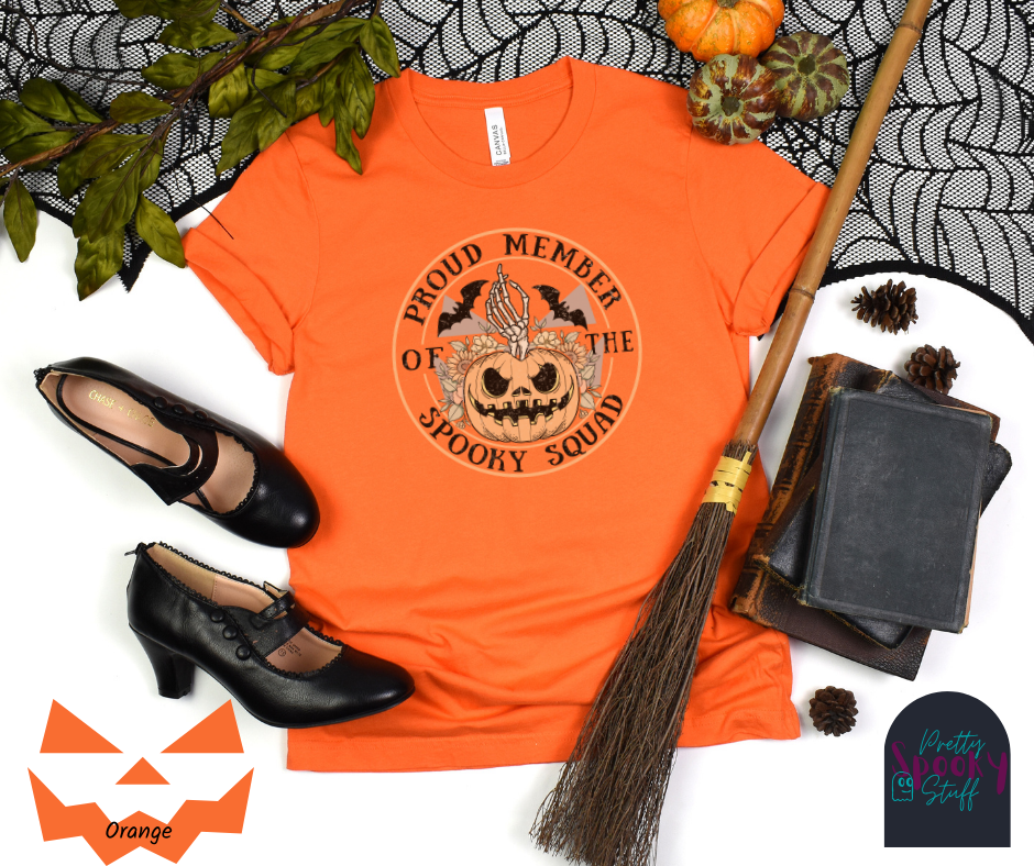 Pumpkin Halloween shirt with jack o' lantern in front of sunflowers, a skeleton hand popping out the top, two bats above, and the words "proud member of the spooky squad" making a circle around it all, faded vintage look, in orange.
