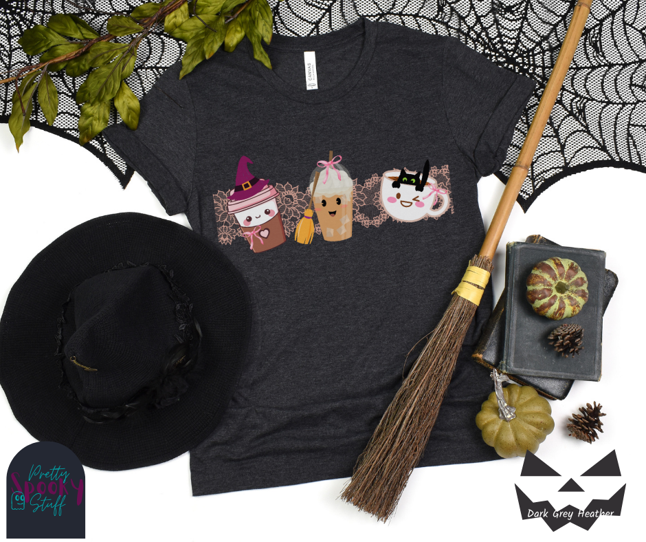 Coquette Halloween shirt with three different coffee cups, a to-go cup wearing a purple witch hat, an iced coffee with a witch broom, and a white mug with black cat hanging on the ledge from the inside, and light pink lace behind them, in dark grey heather.