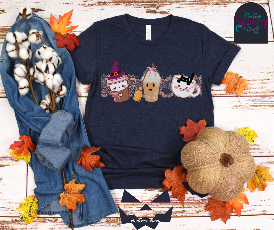 Coquette Halloween shirt with three different coffee cups, a to-go cup wearing a purple witch hat, an iced coffee with a witch broom, and a white mug with black cat hanging on the ledge from the inside, and light pink lace behind them, in heather navy.
