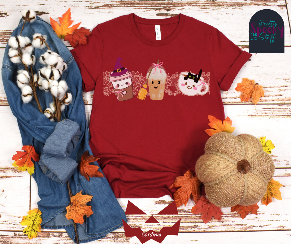 Coquette Halloween shirt with three different coffee cups, a to-go cup wearing a purple witch hat, an iced coffee with a witch broom, and a white mug with black cat hanging on the ledge from the inside, and light pink lace behind them, in cardinal.