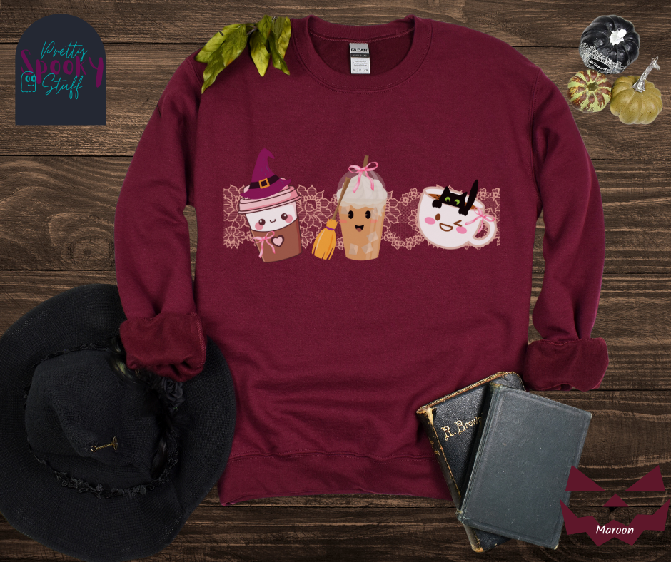 Coquette Halloween sweatshirt with to-go coffee cup character wearing a witch hat and pink bow, an iced coffee cup character with a witch broom and pink bow, and a white coffee mug character with a black cat hanging on to the edge from the inside, with strip of pink intricate lace behind them all, in maroon.