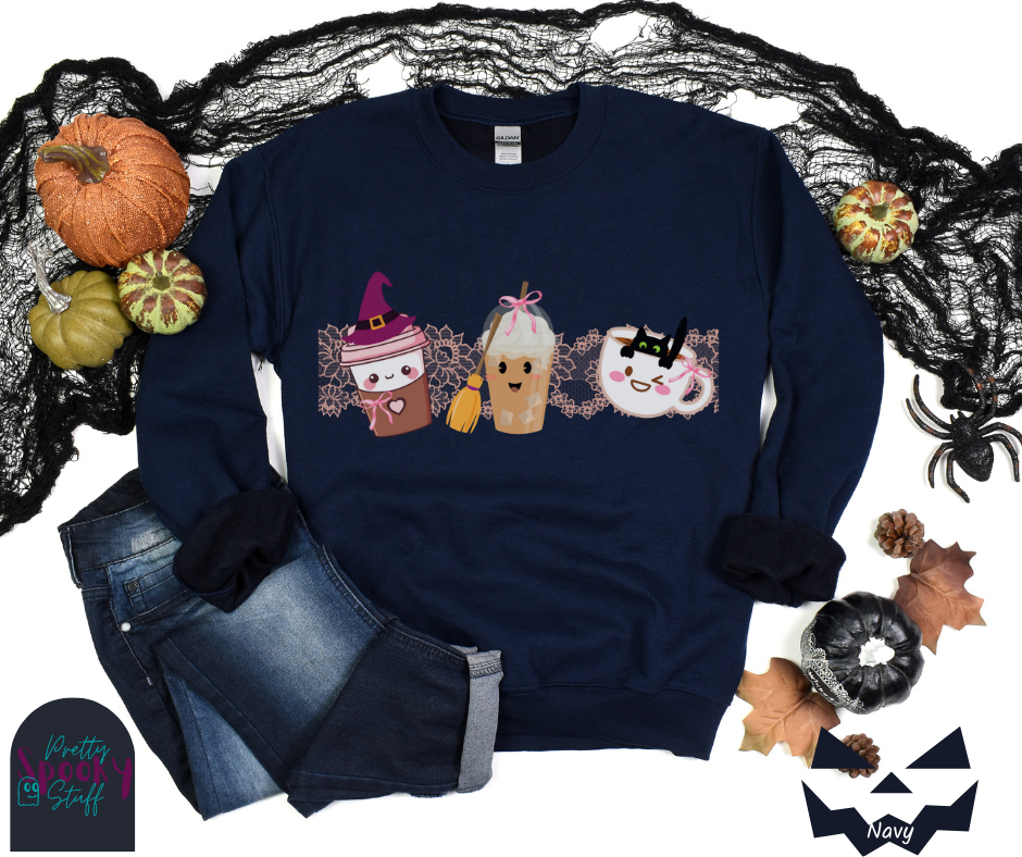 Coquette Halloween sweatshirt with to-go coffee cup character wearing a witch hat and pink bow, an iced coffee cup character with a witch broom and pink bow, and a white coffee mug character with a black cat hanging on to the edge from the inside, with strip of pink intricate lace behind them all, in navy.