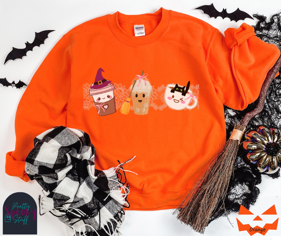 Coquette Halloween sweatshirt with to-go coffee cup character wearing a witch hat and pink bow, an iced coffee cup character with a witch broom and pink bow, and a white coffee mug character with a black cat hanging on to the edge from the inside, with strip of pink intricate lace behind them all, in orange.