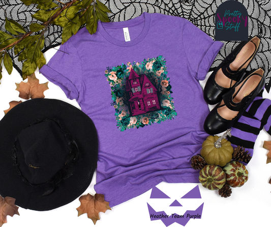 Haunted house Halloween shirt with purple haunted house sitting on top of a rectangle bed of blue and pink flowers, in heather team purple.