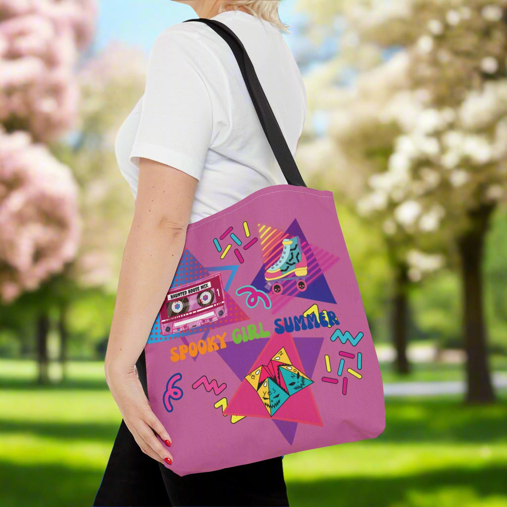 90's Style Summerween tote bag, magenta pink background with pink cassette tape with the words "haunted house mix" on it, rollerskate with bats, and skulls on the wheels, folded paper fortune teller, squiggles, and the words "spooky girl summer" across the middle in orange. green, and dark blue. Woman with bag on her shoulder
