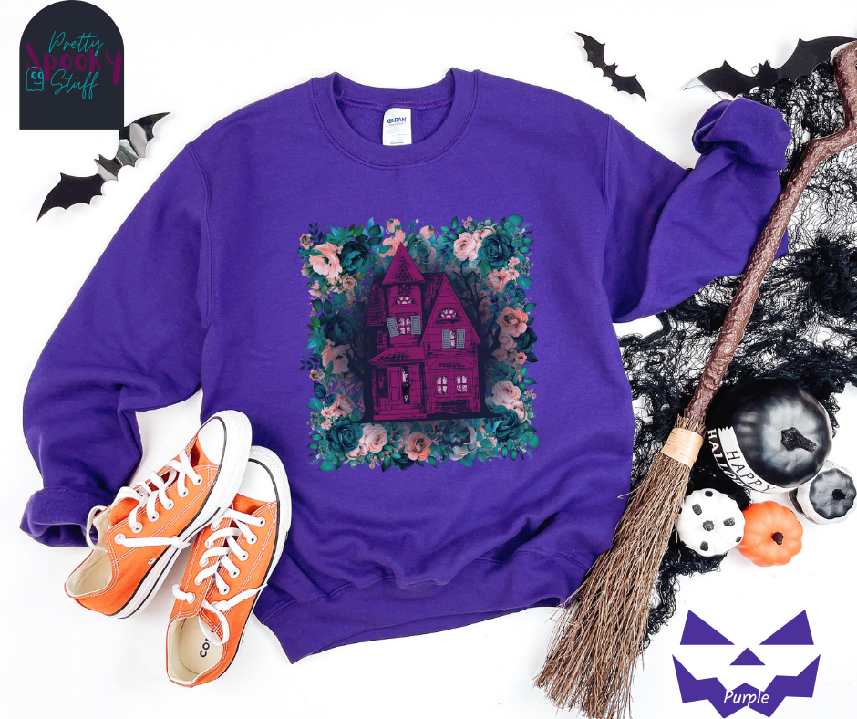 Haunted house Halloween sweatshirt with purple haunted house sitting on top of a rectangle bed of blue and pink flowers, in purple.