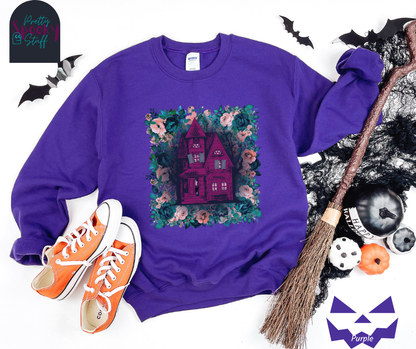 Haunted house Halloween sweatshirt with purple haunted house sitting on top of a rectangle bed of blue and pink flowers, in purple.