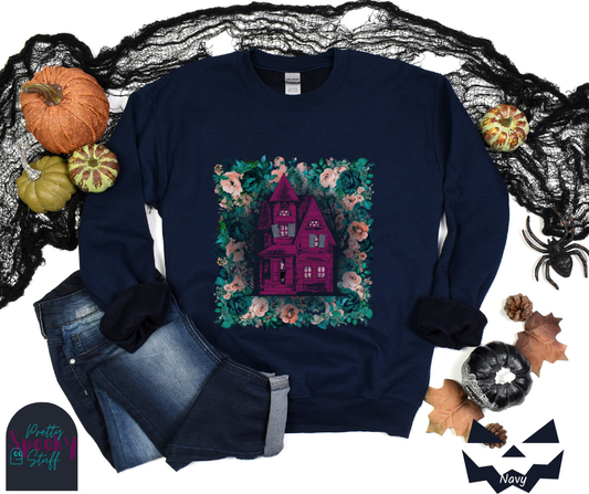 Haunted house Halloween sweatshirt with purple haunted house sitting on top of a rectangle bed of blue and pink flowers, in navy.