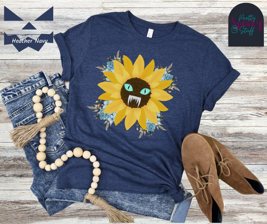 Spooky Girl Summer t-shirt design featuring a sunflower with a menacing face in the middle(striking blue eyes and sharp teeth), with light blue florals in each corner, in heather navy.