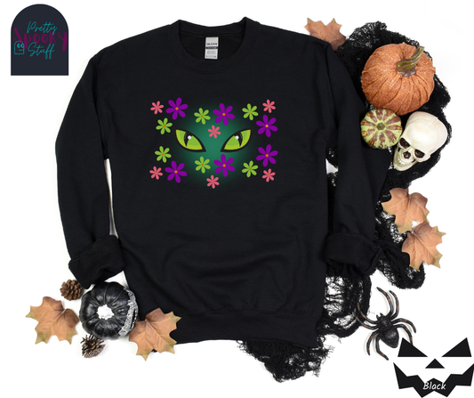Cat Halloween sweatshirt with spooky green cat eyes in the middle of purple, pink and green flowers, with a blue glow behind them, in black.