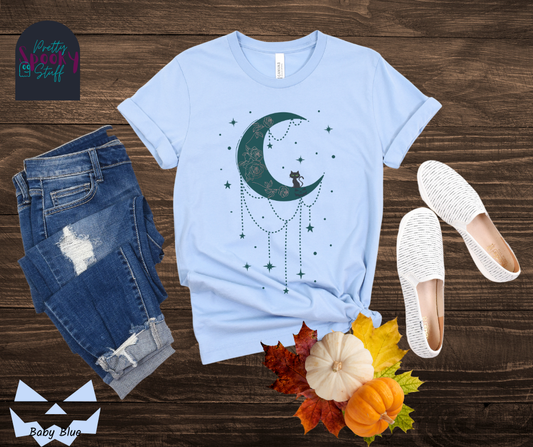 Moon and stars Halloween shirt with a teal crescent moon with pink floral design inside of it, beads hanging from the bottom, a black cat perched on the bottom, and teal stars surrounding it, in baby blue.