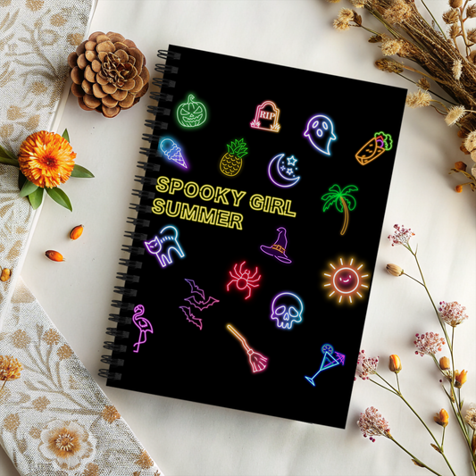 Spooky Girl Summer spiral notebook featuring a mix of spooky and summer themed neon icons with the with the words "spooky girl summer" in neon yellow letters in the middle, black background, sitting on decorated table.