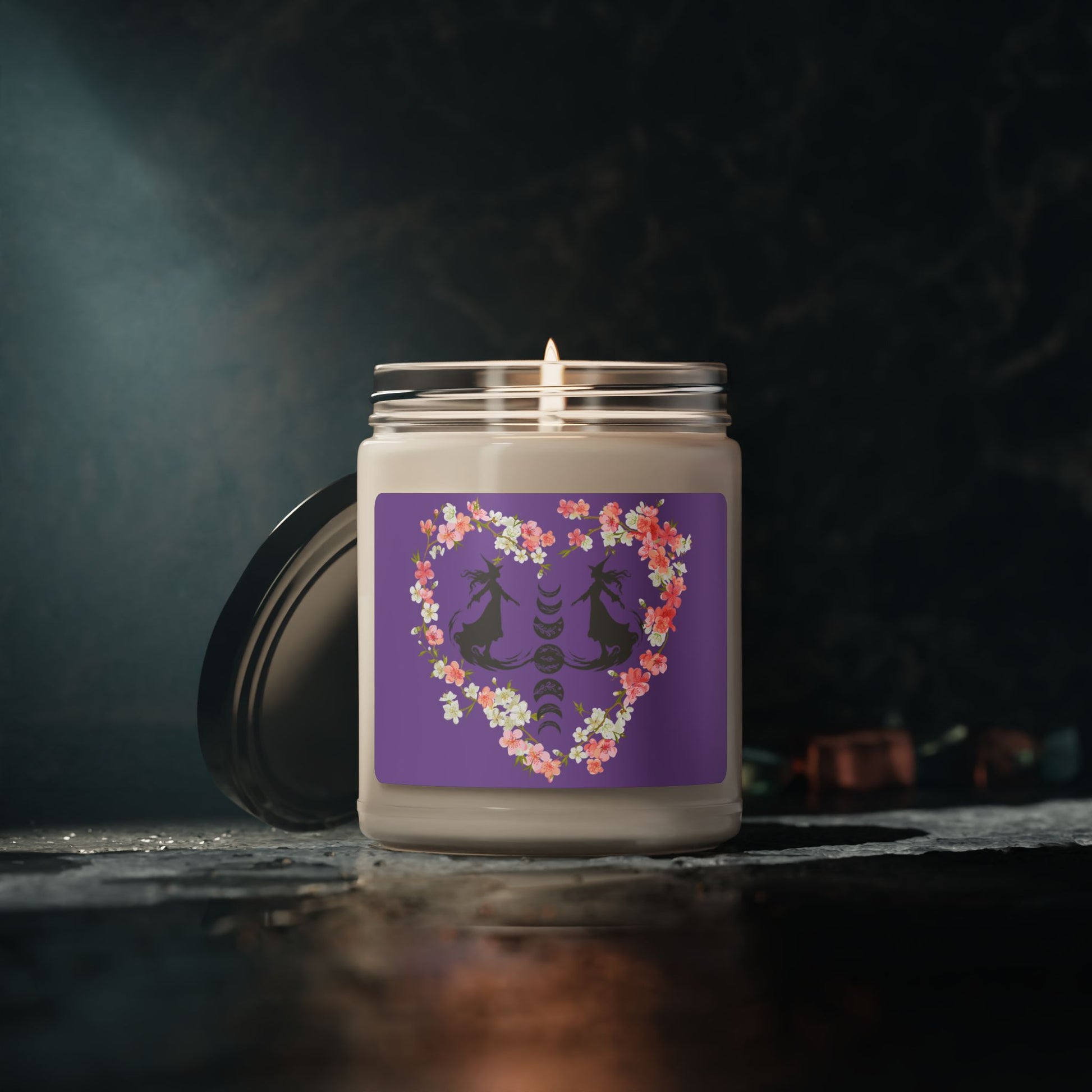 Witch themed summerween soy candle featuring the silhouettes of two graceful witches facing each other in mid glide, the phases of the moon between them with floral details, surrounded by a heart made from pink and white cherry blossoms, with a medium purple sticker background, sitting on a dark table with dark marble wall.