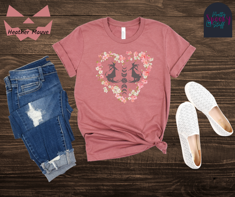 Witch themed summerween shirt with silhouettes of two graceful witches mid-step on what seems like fog, with the moon phases going top to bottom in between them, surrounded by a heart of pink and white cherry blossoms, in heather mauve.