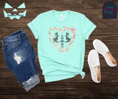 Witch themed summerween shirt with silhouettes of two graceful witches mid-step on what seems like fog, with the moon phases going top to bottom in between them, surrounded by a heart of pink and white cherry blossoms, in heather mint.