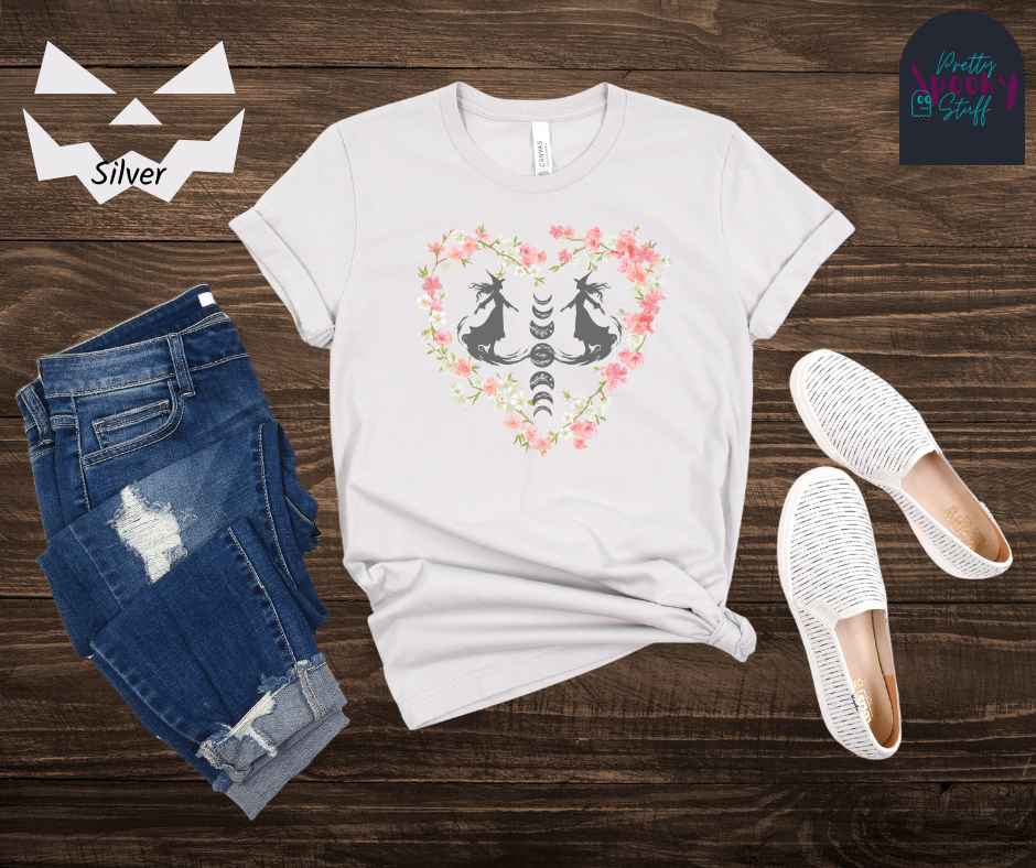 Witch themed summerween shirt with silhouettes of two graceful witches mid-step on what seems like fog, with the moon phases going top to bottom in between them, surrounded by a heart of pink and white cherry blossoms, in silver.