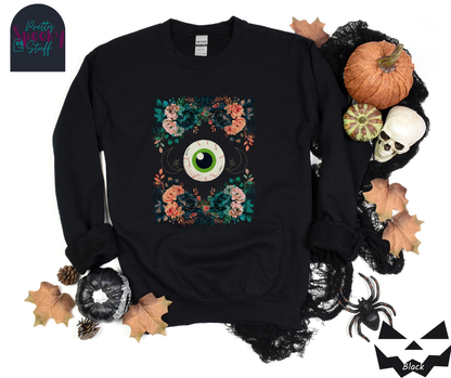 Floral Halloween sweatshirt with a large eyeball in the middle, gold crescent moon and star line drawings on either side, surrounded by four bunches of flowers that make a frame, in black.