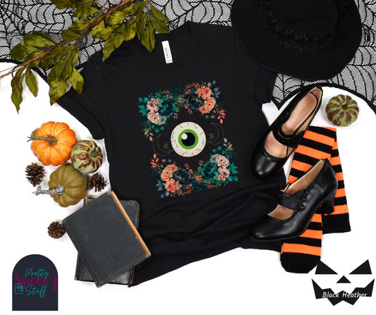Unique Halloween shirt with large eye in the middle, with gold line drawing of crescent moon and star, and four bunches of pink and blue flowers forming a frame around it, in black heather.