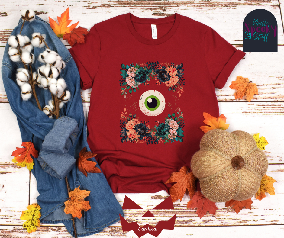 Unique Halloween shirt with large eye in the middle, with gold line drawing of crescent moon and star, and four bunches of pink and blue flowers forming a frame around it, in cardinal.