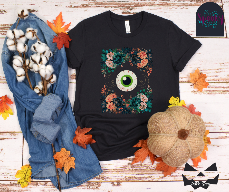 Unique Halloween shirt with large eye in the middle, with gold line drawing of crescent moon and star, and four bunches of pink and blue flowers forming a frame around it, in dark grey.
