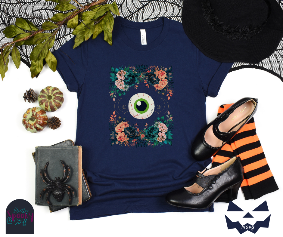 Unique Halloween shirt with large eye in the middle, with gold line drawing of crescent moon and star, and four bunches of pink and blue flowers forming a frame around it, in navy.