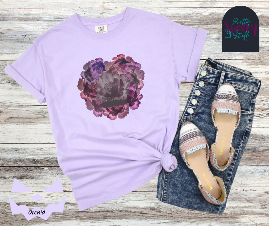 Summerween shirt with flowers of different shades of purple arranged in the shape of a heart, with shadows of running zombies and two zombie arm line drawings mixed in, in orchid.