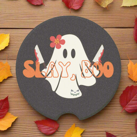 Halloween car coaster with a ghost wearing a pink flower, holding two bloody knives with the words "slay boo" in orange vintage font in front, black background.