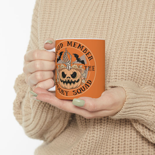 Halloween ceramic mug with a skeleton hand coming out of a jack o lantern with flowers behind it, two bats above it, surrounded by the words "proud member of the spooky squad" in a circle, orange background.