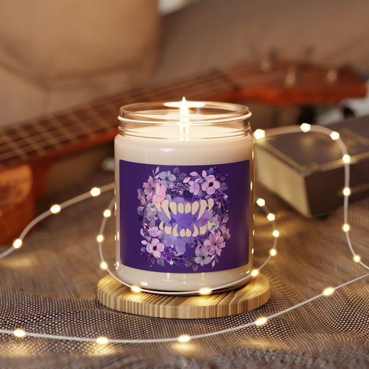 Spooky girl summer 9oz soy candle featuring a label with florals in shades of purple, with a set of vampire teeth in the middle, and a pink butterfly in a subtle perch on a fang, on dark purple background. Sitting on a table with fairy lights.