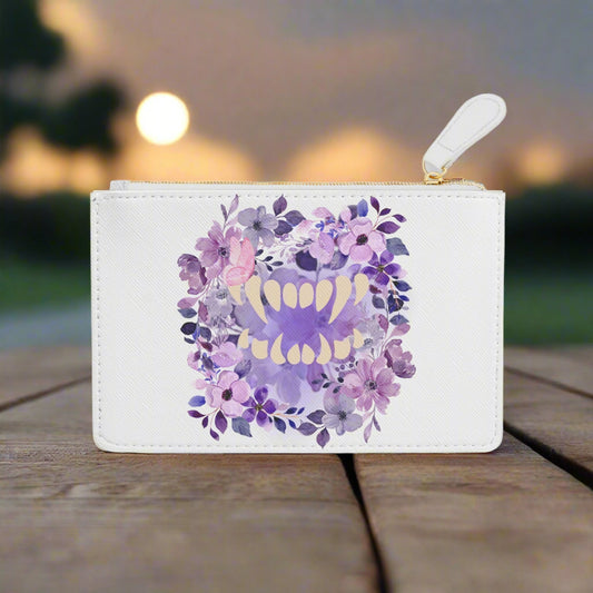 Spooky girl summer mini clutch bag featuring a bed of florals in shades of purple, with a set of vampire teeth on top, and a pink butterfly in a subtle perch on a fang, on white faux leather material. Sitting on a wooden surface.