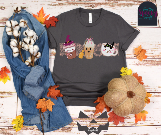 Coquette Halloween shirt with three different coffee cups, a to-go cup wearing a purple witch hat, an iced coffee with a witch broom, and a white mug with black cat hanging on the ledge from the inside, and light pink lace behind them, in asphalt.