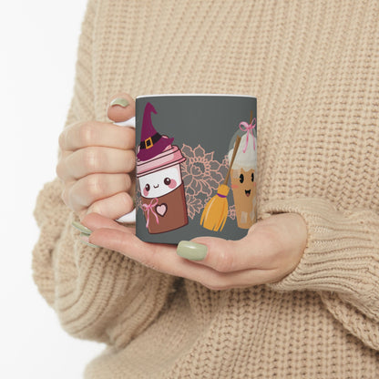 Coquette Halloween 11oz ceramic mug with three different coffee cups, a to-go cup wearing a purple witch hat, an iced coffee cup with a witch broom, and a white mug with a black cat hanging on the edge from the inside, with a light pink lace strip behind them, gray background, two hands holding it against a sweater.