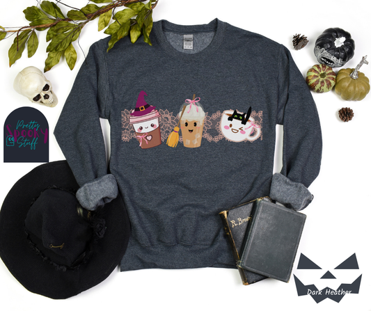 Coquette Halloween sweatshirt with to-go coffee cup character wearing a witch hat and pink bow, an iced coffee cup character with a witch broom and pink bow, and a white coffee mug character with a black cat hanging on to the edge from the inside, with strip of pink intricate lace behind them all, in dark heather.